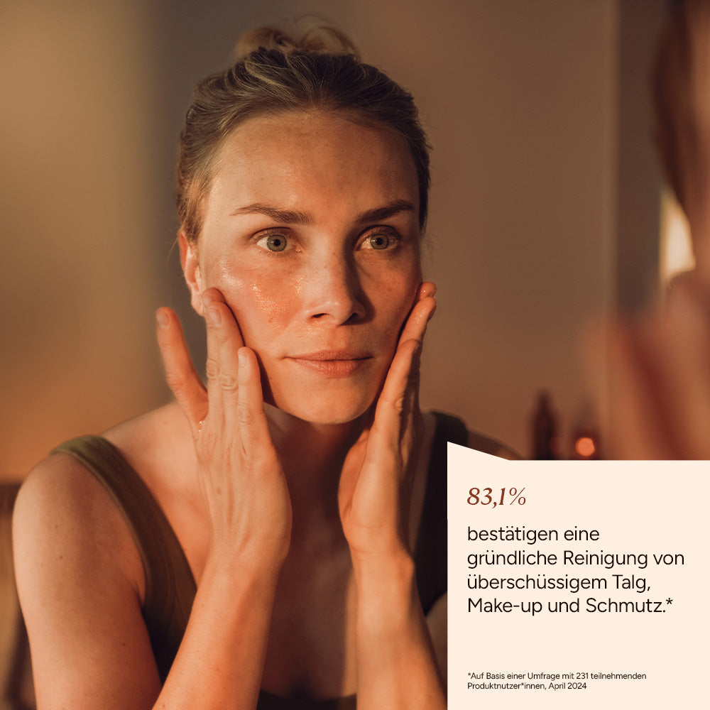 Eine Frau trägt Reinigungsoel auf ihr Gesicht auf, sieht in den Spiegel. Die Atmosphäre ist warm und entspannt.