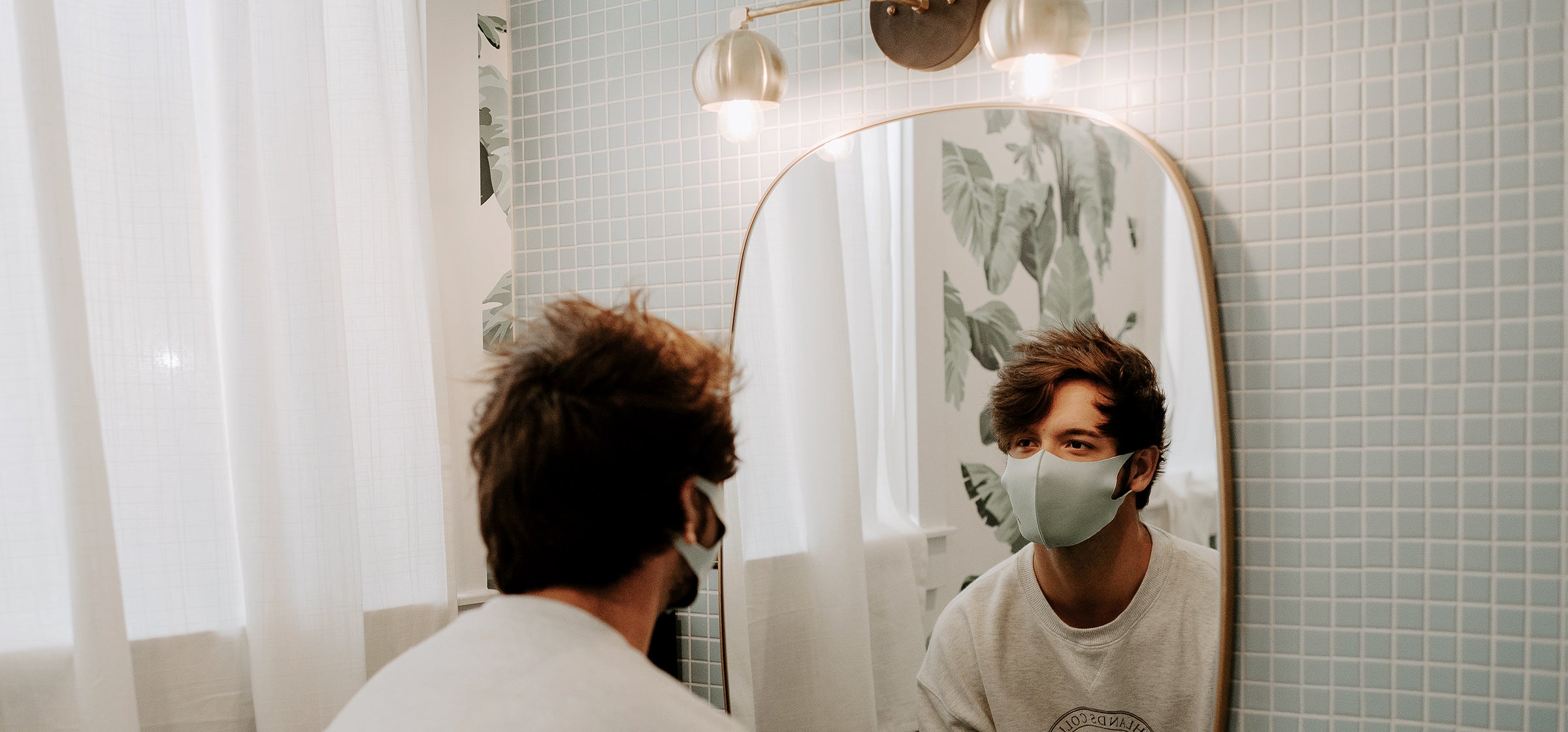 Man stands in a bathroom and looks at himself in the mirror. He is wearing a face mask.