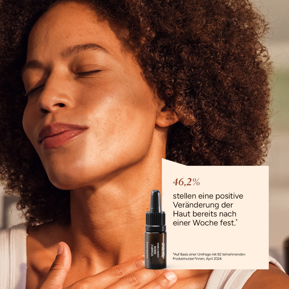 Een vrouw met krullend haar streelt zachtjes haar huid; op de voorgrond staat een bruine glazen fles met de tekst ‘ Vitamine C Serum ’.
